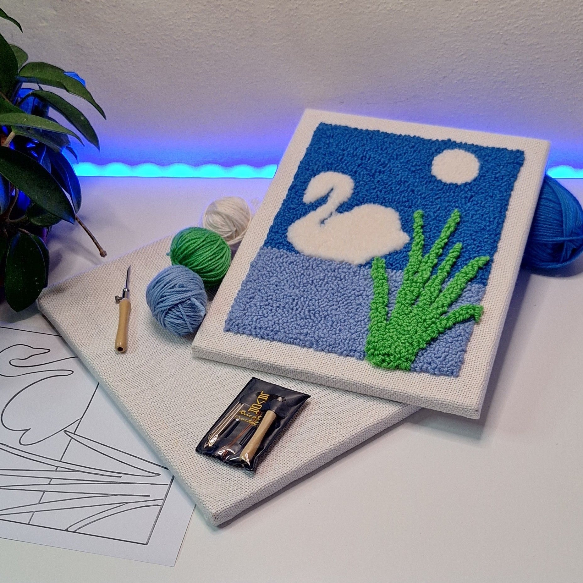 quadro ricamato con punch needle che raffigura un cigno bianco sull'acqua azzurra e cielo blu. Si vedono l'ago utilizzato per il ricamo sia fuori che dentro la sua confezione, i gomitoli e il telaio da ricamo.