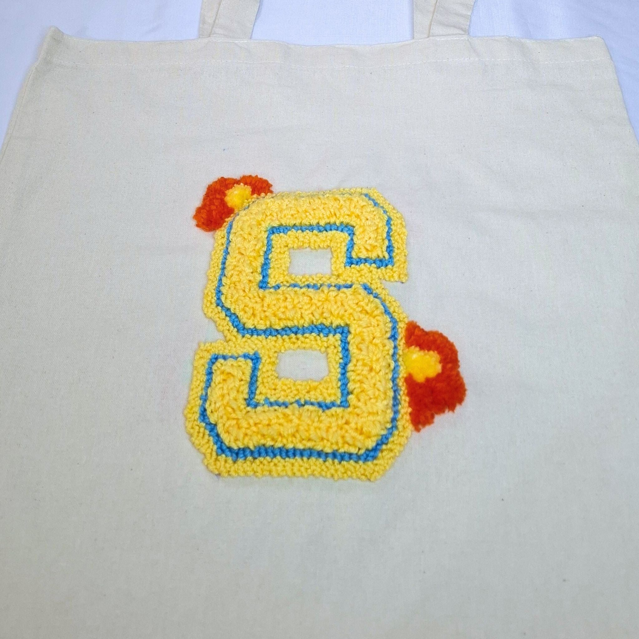ingrandimento del ricamo della lettera S con punch needle. La lettera è di colore giallo con un contorno blu e due fiorellini arancioni/gialli come ornamento
