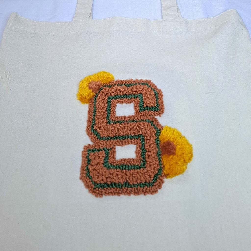 ingrandimento del ricamo della lettera S con punch needle. La lettera è di colore rosso mattone/marrone con un contorno verde e due fiorellini marron/gialli come ornamento