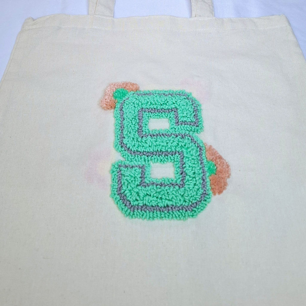 ingrandimento del ricamo della lettera S con punch needle. La lettera è di colore verde smeraldo con un contorno grigio e due fiorellini rosa/verdi come ornamento