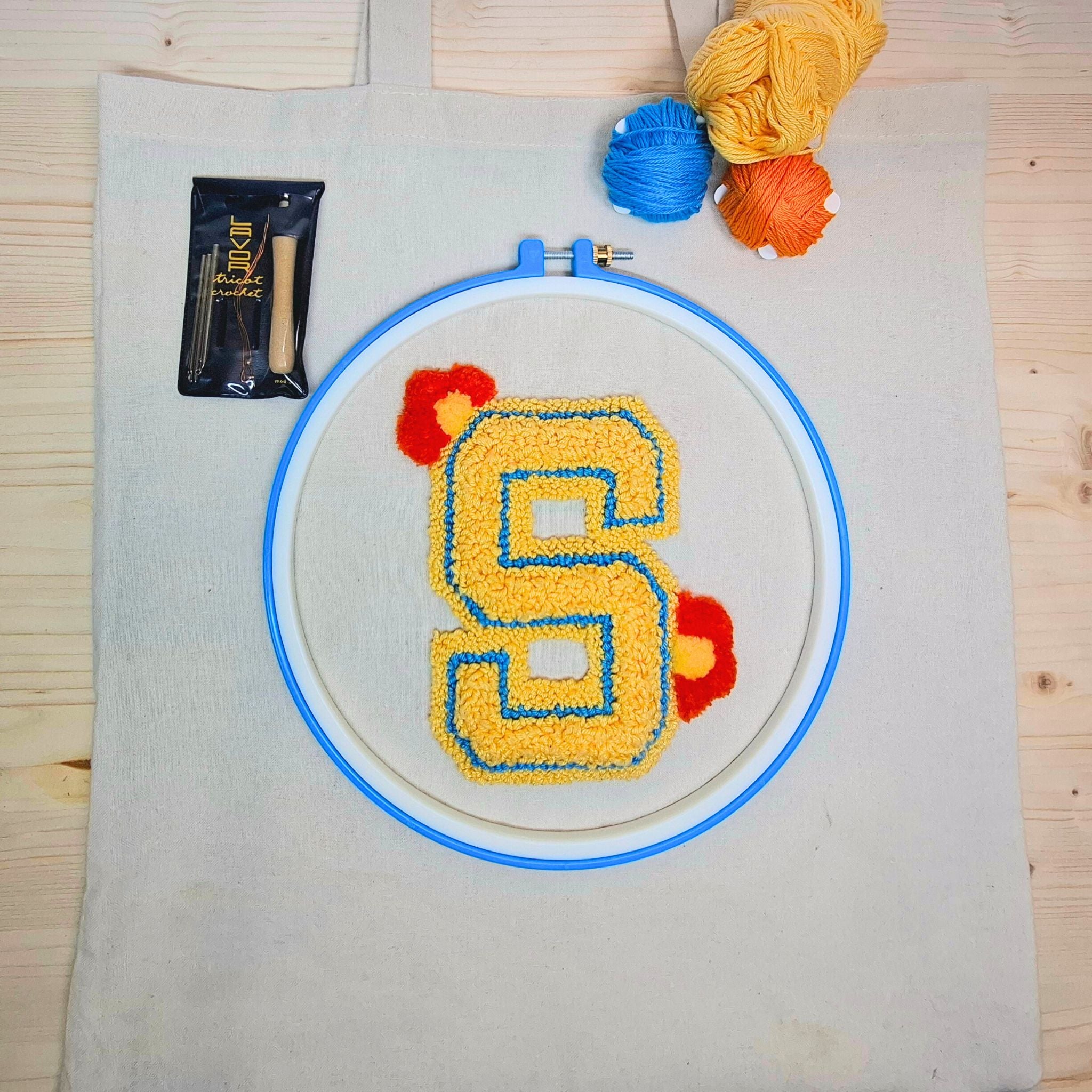 borsa shopper stesa su un tavolo di legno chiaro con un ricamo in punch needle sulla parte centrale che raffigura la lettera S gialla. Si vedono tutti i componenti del kit da ricamo.