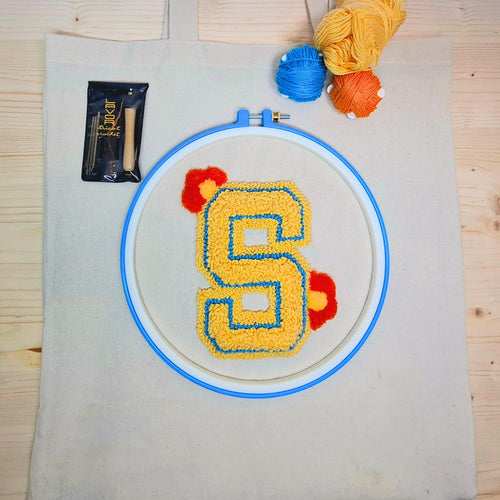borsa shopper stesa su un tavolo di legno chiaro con un ricamo in punch needle sulla parte centrale che raffigura la lettera S gialla. Si vedono tutti i componenti del kit da ricamo.